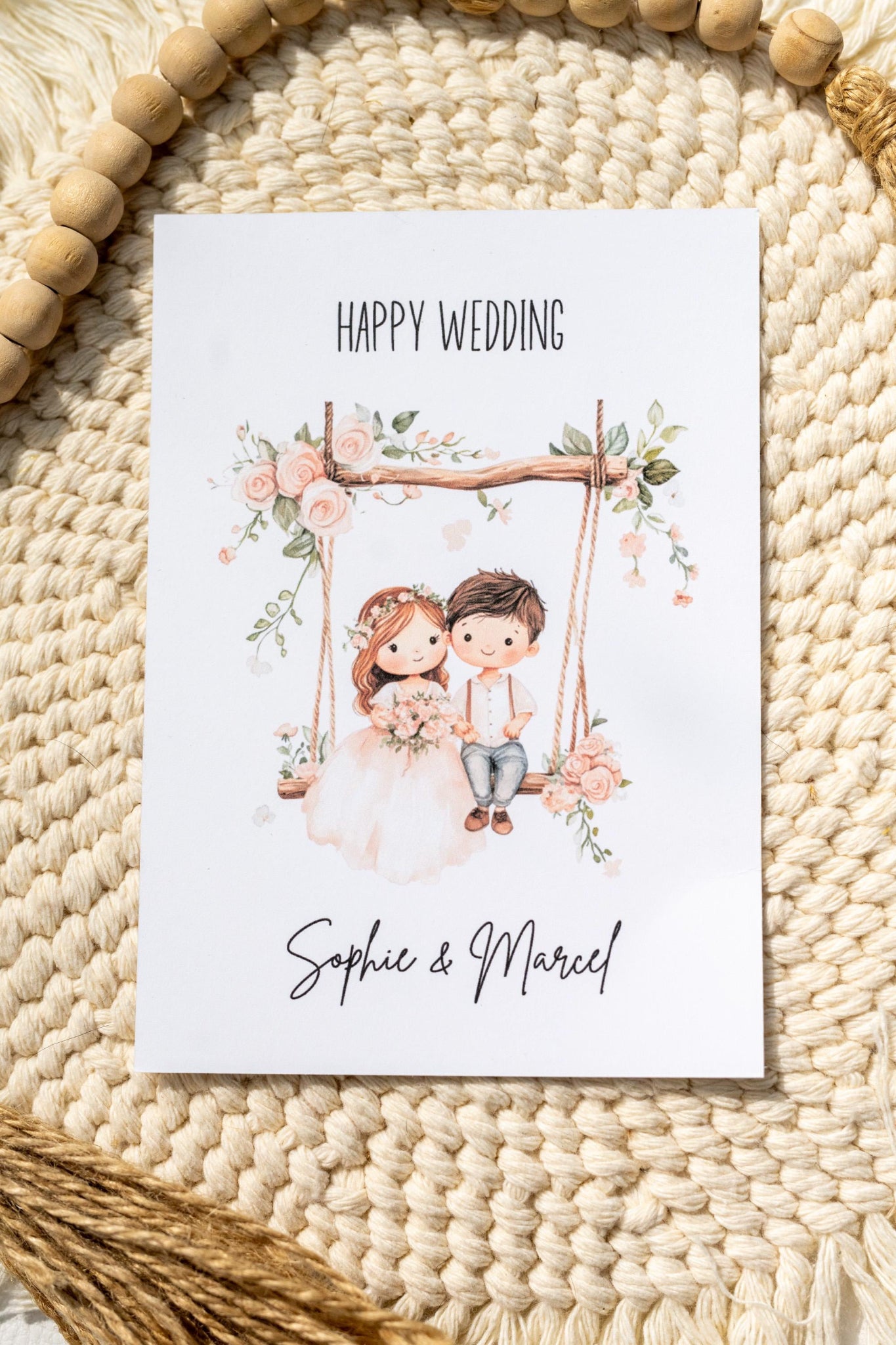 Süße Karte zur Hochzeit mit einem Brautpaar auf einer Schaukel mit Blumen im Aquarell-Look. Der Text oben auf der Karte lautet Happy Wedding und unten stehen die Vornamen des Brautpaars. Das Format der Karte ist DIN A6.